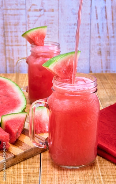 Fototapeta splash of watermelon water in a glass jar with a watermelon triangle on top on a wooden table and white wooden plank background.