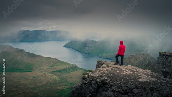 Fototapeta Man in red jacket overlooking green mountain panorama with view to fjord from highest mountain summit on Faroe Islands, Slættaratindur on island Eysturoy.