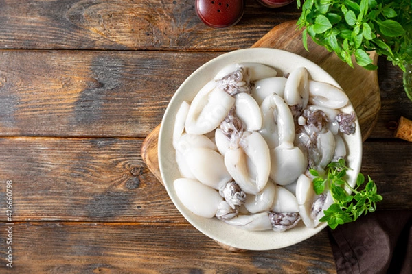 Obraz Raw cuttlefish in a ceramic plate on the kitchen table. Lots of peeled squid in a bowl on a brown wooden table top view. The concept of cooking shellfish