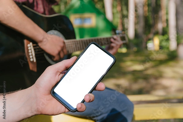 Fototapeta Mock up of a smartphone in front of a man playing the guitar. Against the background of the forest and houses in nature.