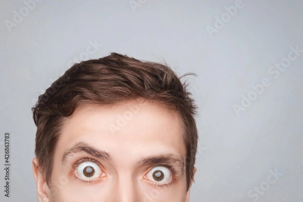 Fototapeta Man's eyes looking up on the light background. Empty place for a text or object. Close-up shot of shocked young man with round eyes, top half head. Eyes looking on front