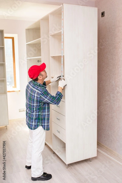 Fototapeta The furniture assembler measures the height of the cabinet with a construction tape during assembly