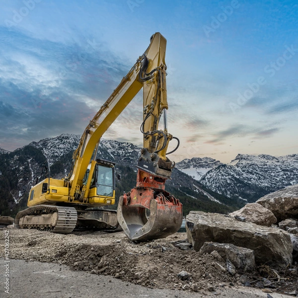 Obraz yellow chain excavator with grapple of natural stone in front of a mountain backdrop and cloudy sky