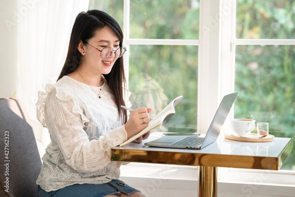 Fototapeta Happy of asian freelance people Businesswoman Taking written Notes to on notebook casual working with laptop computer with a coffee cup mug and smartphone at the cafe,Business Lifestyle
