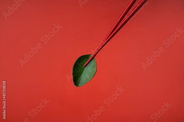 Obraz Derazht green lime leaf with red Chinese chopsticks. Red on red. Cooking ingredients for Asian food. Spicy food. Red background. Asian products. Soup ingredient tom yang. Macro photography of food.