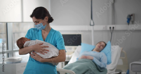 Fototapeta Nurse in medical mask holding newborn baby with mother resting in bed on background