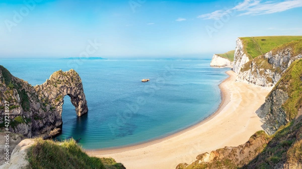 Fototapeta Aerial view of Durdle Door natural formation at UNESCO heritage Jurassic Coast. The Isle of Portland can be seen on the horizon. Copy space in blue sky.