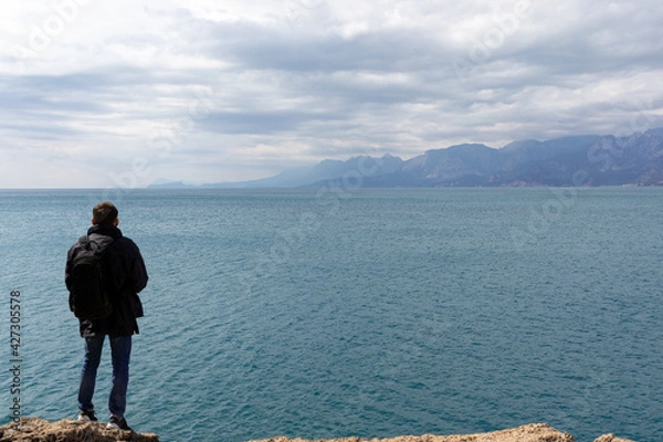 Fototapeta Young casual man traveller with backpack stands on high stone shore and looking in the sea distance. Web banner for travel and exploring the natural world.
