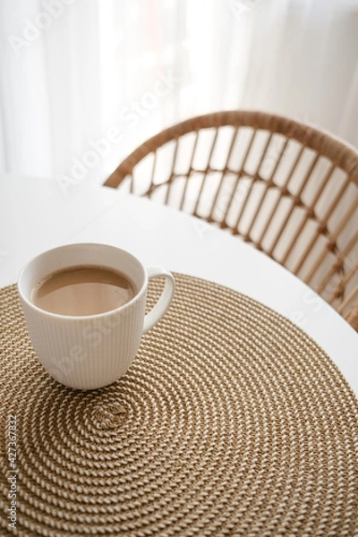 Fototapeta White cup of coffee on wicker place mat. Background with white table and rattan chair. Boho style kitchen. Copy space. Hygge.