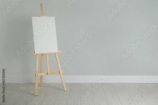 Fototapeta Wooden easel with blank canvas near light wall. Space for text