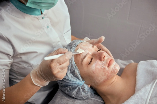 Fototapeta A female cosmetologist in a medical mask applies cream to the client's face with a brush. Hydration and rejuvenation of the skin. Professional skin care.
