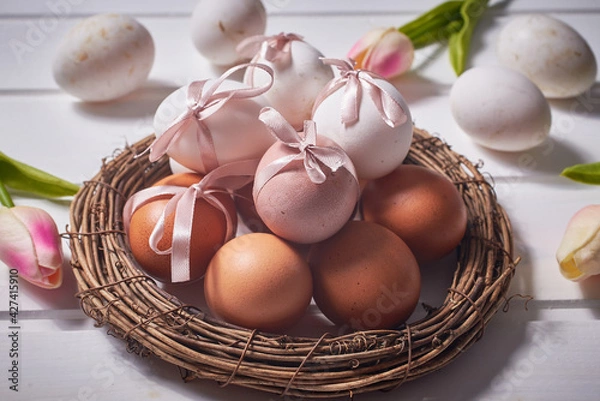 Fototapeta White and brown eggs with pink bows on a nest on a white wooden background and tulip flowers.