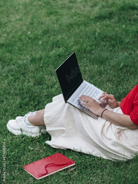 Obraz A girl in a red shirt and white skirt is sitting on the grass with a laptop. Study in the fresh air. Remote work in nature. Working on a laptop. The girl has light skin. No face visible.