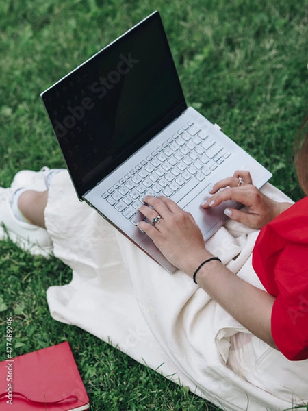 Obraz A girl in a red shirt and white skirt is sitting on the grass with a laptop and typing. Study in the fresh air. Remote work in nature. Working on a laptop. The girl has light skin. No face visible.