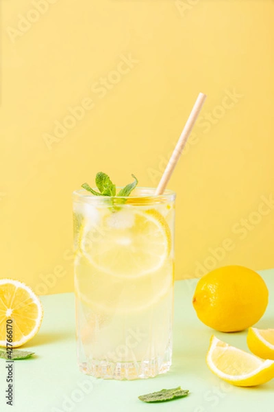 Fototapeta Tall glass of ice tea or water with ice cubes, lemon and mint leaves on a combined colored yellow and green background. Cool refreshing carbonated cocktail for summer time