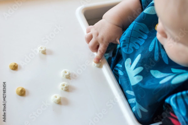 Fototapeta Baby using pointer finger to drag a cereal puff to edge of tray: baby led weaning made fun with smiley face made of cereal puffs