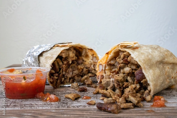 Fototapeta A carne asada burrito cut in half with meat falling out onto the table next to a cup of salsa.