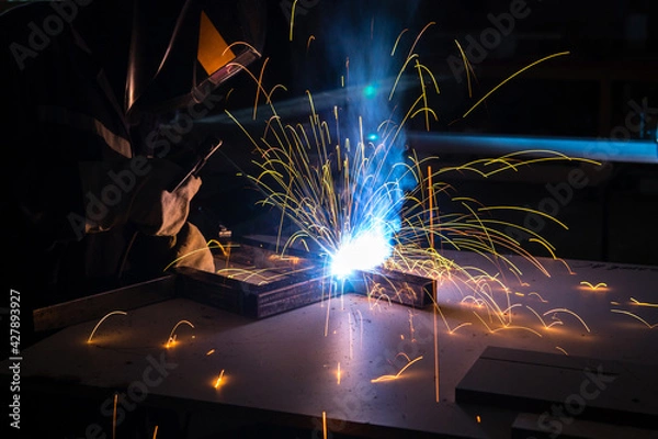 Fototapeta A man welder with construction gloves and a welding mask is welded with a welding machine metal in workshop.