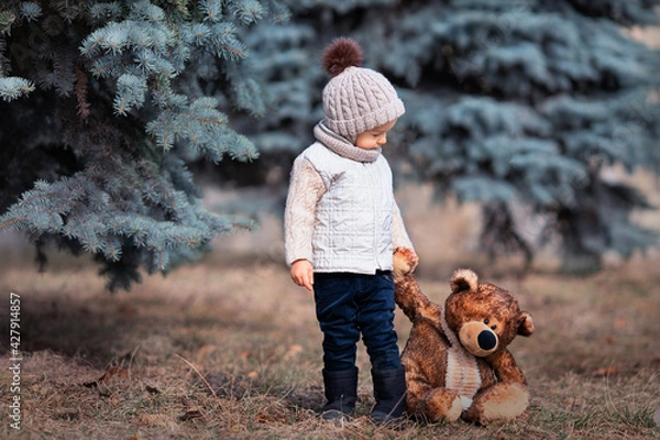 Fototapeta Baby and his friend bear in the park. Winnie the Pooh. Spring walk of a child with a toy. Based on a fairy tale. Brown teddy bear and a boy in a knitted hat and jacket