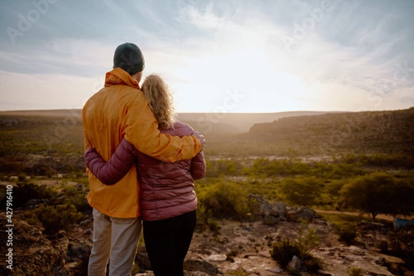 Obraz Rear view of young couple in love embracing while looking at beautiful sunrise after hiking to mountain cliff edge