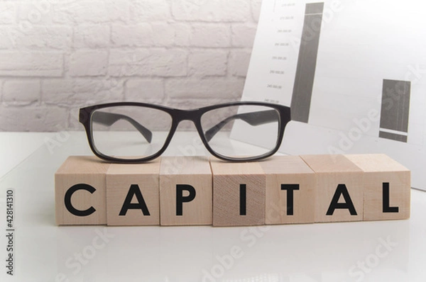 Fototapeta The word CAPITAL is written on wooden cubes standing on the white surface of the table, the GLASSES are on the cubes