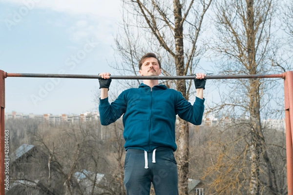 Obraz Young man in sportswear exercising outside, pulling himself up on a horizontal bar. Power workout in the fresh air