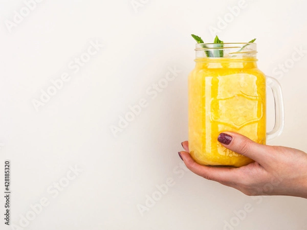 Obraz Woman hand holding orange smoothie shake with mint in the glass jar against beige wall.