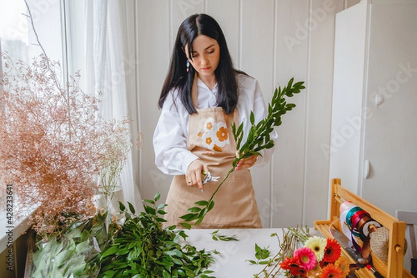 Fototapeta a young brunette woman florist in apron cuts leaves from the stems before starting to create a bouquet in her workshop