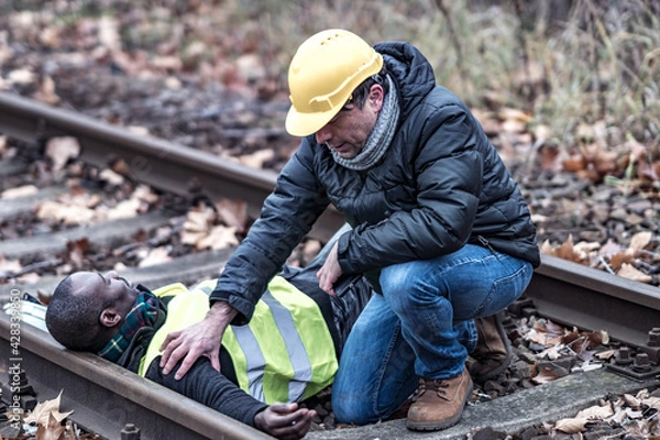 Obraz African American railroad engineer injured in an accident at work on the railway tracks. Coworker helping him
