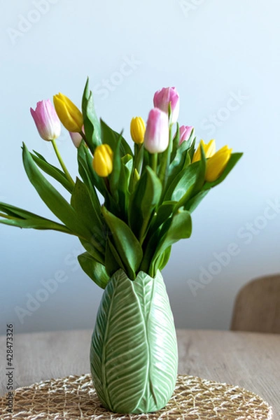Obraz Vertical photo of a beautiful composition of pink and yellow tulips inside a green vase on a wooden table. White background and selective focus.