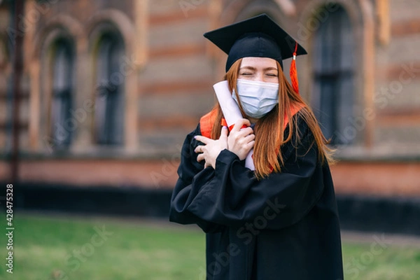 Obraz A female graduate student in a protective medical mask, in a black graduation dress, with a diploma in her hands. Graduation ceremony concept, quarantine, coronavirus