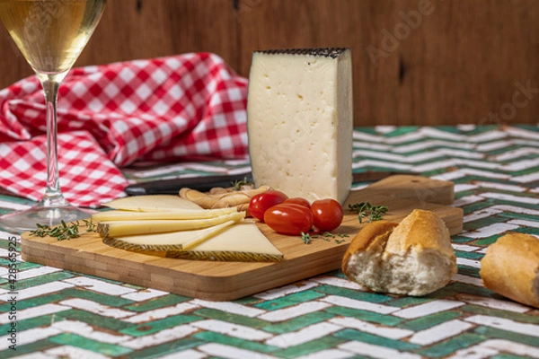 Fototapeta Spanish Manchego cheese sliced with some cherry tomatoes and a glass of white wine over a green and white table.
