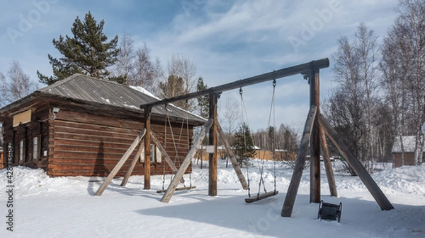 Fototapeta The swing is made of logs and is suspended from ropes. Nearby there is an unpainted wooden one-story house. Snow on the ground. Sunny winter day