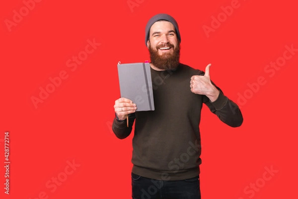 Fototapeta Portrait of smiling hipster bearded man holding planner and showing thumb up