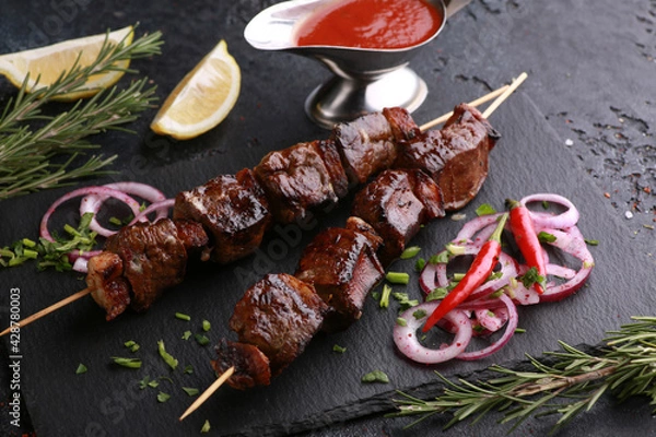 Fototapeta Fried liver shish kebab with lard on skewers with rosemary, lemon, fresh vegetables, herbs and tomato sauce on black table. Background image, copy space