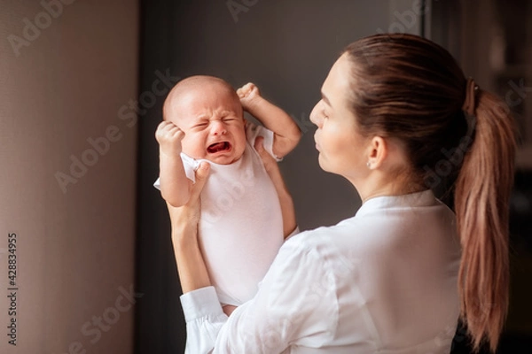 Obraz Mother with tail in white shirt holding crying newborn baby. Copy space.