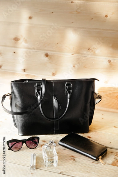 Fototapeta black rectangular leather bag with a rectangular leather wallet and sunglasses on a wooden table in the room. sunny day