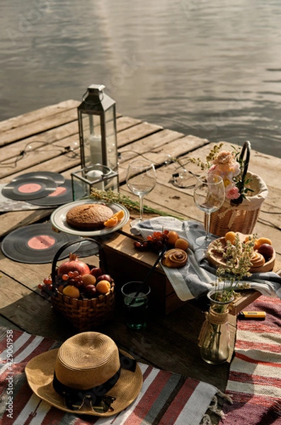 Fototapeta Romantic picnic by the river with fresh fruit, pastries and wine. A romantic outdoor dining experience and the beauty of the setting sun. Vertical orientation.