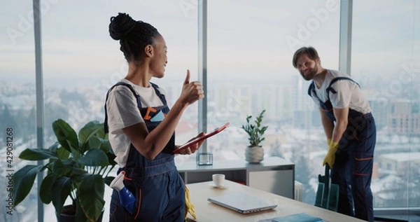 Obraz Cleaning services team. Young man using vacuum cleaner tidying office workspace. Female african american cleaning employee using tablet computer.