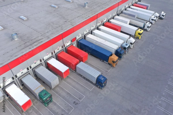 Fototapeta Aerial view of goods warehouse. Logistics center in industrial city zone from above. Aerial view of trucks loading at logistic center. View from drone.