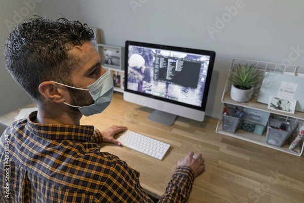 Obraz man works from home with his computer, dark-haired man in a mask and a plaid shirt writes on his home computer on a wooden desk