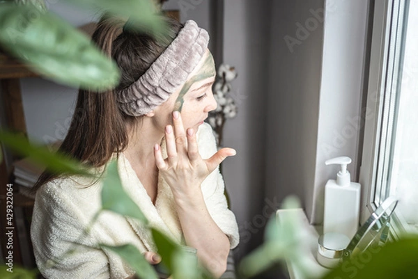 Obraz Young pretty woman in a bathrobe in front of a mirror is applying a green cosmetic mask to her face. Concept of skin care, the use of natural cosmetics and lifestyle
