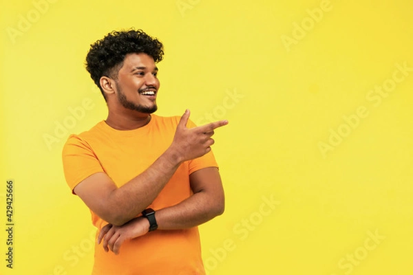 Fototapeta Cheerful smiling man points to a place for text on a yellow background, he is wearing an orange T-shirt, good news. Indian man