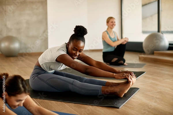 Obraz Happy black athletic woman doing stretching exercise while practicing Pilates with group of women in health club.