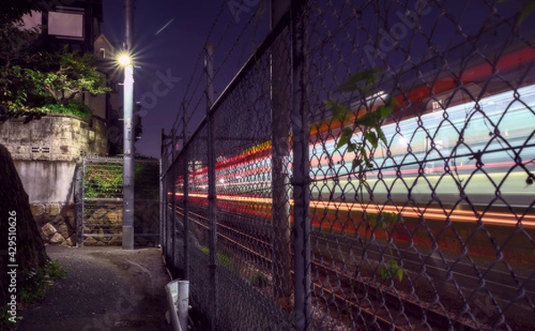 Obraz Long-exposure capture of light trails made by a train passing by an alleyway, separated from the tracks by a chain link fence