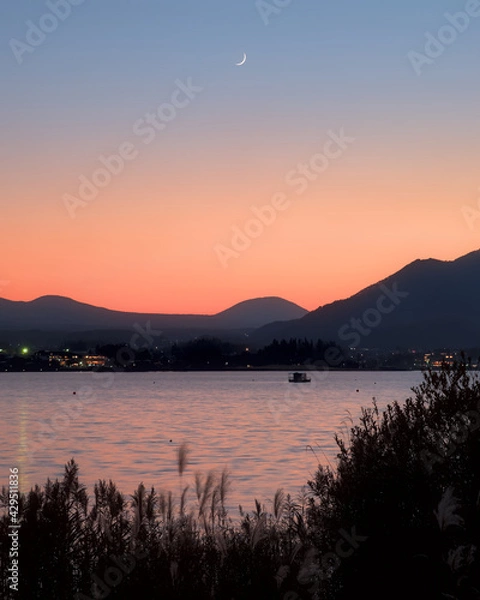 Obraz A crescent moon rising over Lake Kawaguchi and a blue and orange sky at dusk, with low mountains and a boat visible