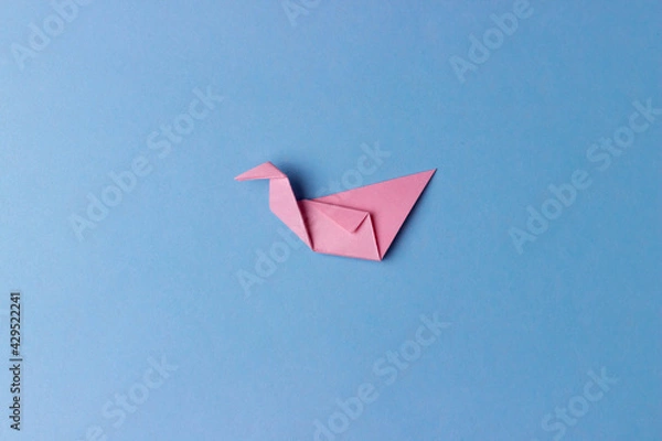 Fototapeta An origami bird folded from pink paper on a blue background in the center of the photo. Education, pastime, hobbies, activities with children. Minimalistic background