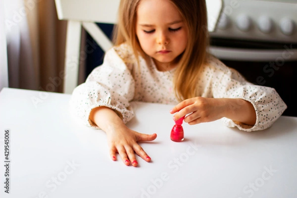 Fototapeta Little girl paints her nails with pink nail polish. Cute toddler girl playing with colorful nail polish doing manicure. Cosmetics and beauty concept.