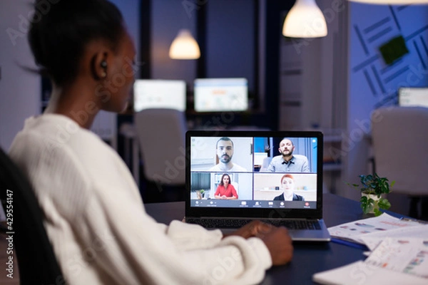 Fototapeta African employee working overtime from business office late at night discussing with partners online using webcam. Diverse team talking during corporate meeting using technology network wireless.