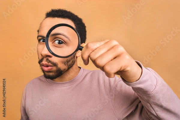 Fototapeta Curious young african american black man with magnifying glass Isolated over beige background. Surprised young man student holding magnifying glass.